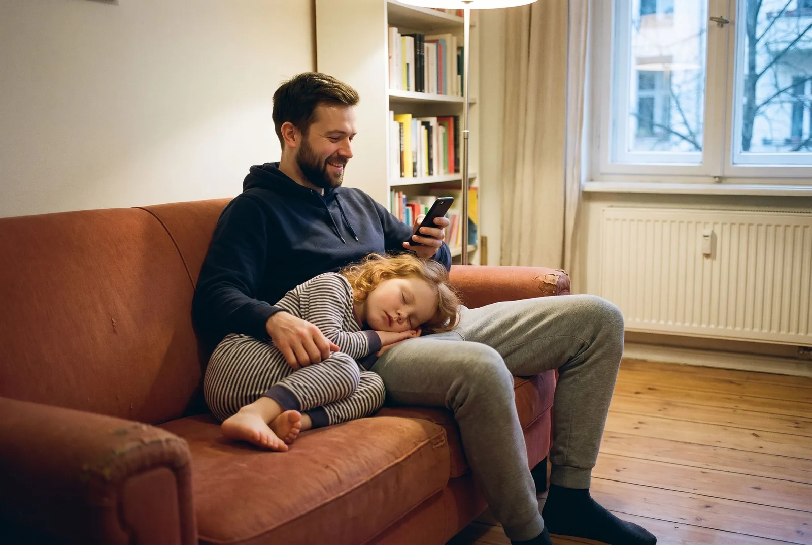 Ein Vater sitzt abends mit dem Handy auf dem Sofa, seine Tochter ist an ihn gelehnt eingeschlafen